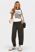 A woman modeling a white ringer t-shirt with a black graphic of a leopard and the text 'WILD HEART', paired with dark gray leopard-print pants, white sneakers, and a black shoulder bag, against a light gray background.