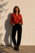 Woman wearing a red patterned blouse and black pants against a white wall.
