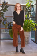Woman in black top and brown pants standing indoors with plants in the background
