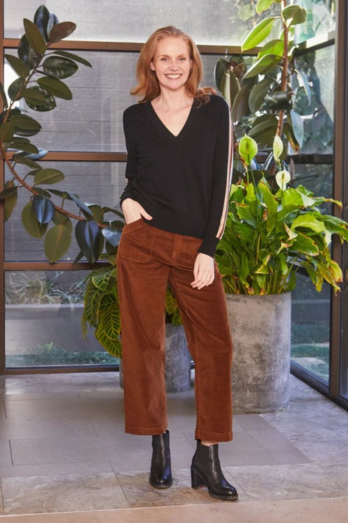 Woman in black top and brown pants standing indoors with plants in the background