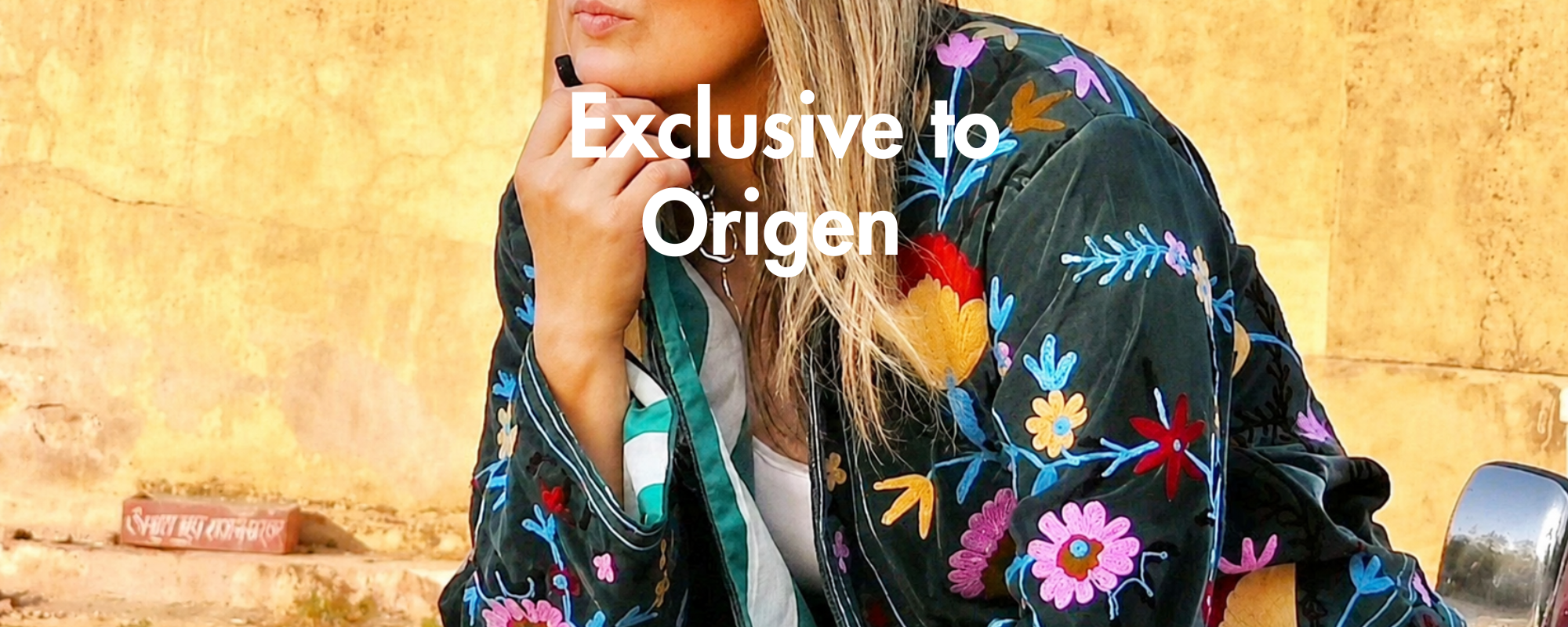 A woman wearing a black velvet jacket with colorful floral embroidery, posed against a textured yellow wall. White text overlay reads 'Exclusive to Origen'.