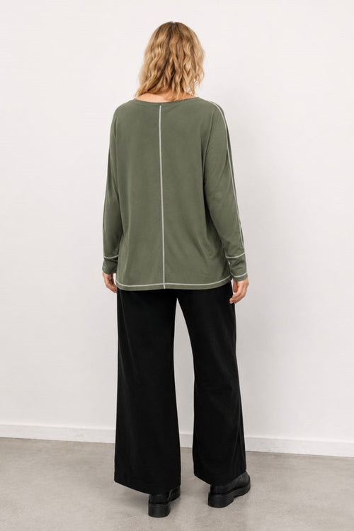 Person wearing a green long-sleeve top and black pants against a white background