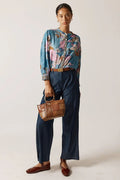 Person wearing a colorful floral blouse and navy pants, holding a brown handbag on a plain background