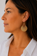 A close-up side profile of a woman wearing a large, gold-toned, fan-shaped dangle earring with a textured, ribbed pattern.
