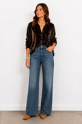 Woman wearing a brown velvet shirt and blue jeans against a white wall.