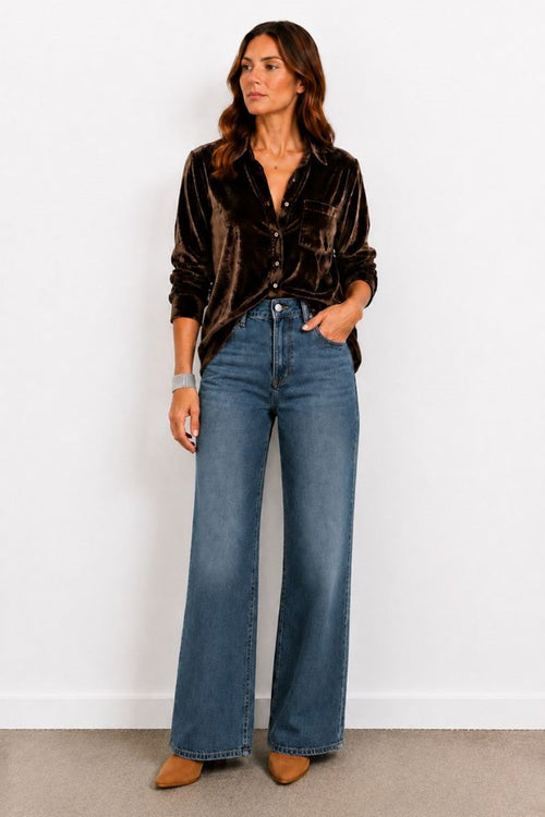 Woman wearing a brown velvet shirt and blue jeans against a white wall.