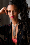 A close-up portrait of a woman with long dark braids wearing a silver rhinestone choker necklace with a long fringe tassel, a black floral robe, and a pink top.