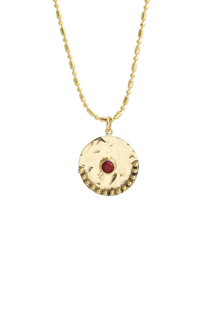 A gold-tone necklace featuring a textured circular pendant with a small red gemstone in the center and a beaded border, shown against a plain white background.