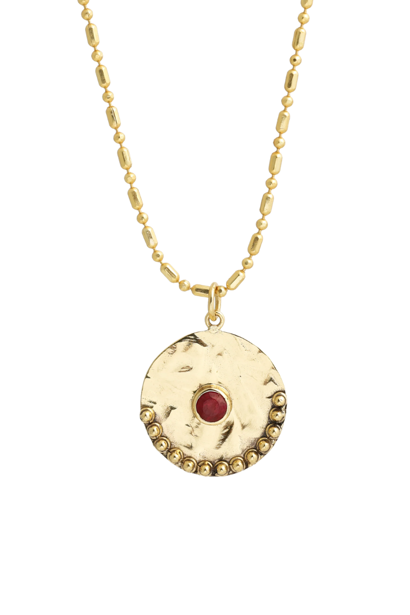 A gold-toned necklace featuring a textured circular pendant with a small red gemstone in the center and a beaded gold border, shown against a plain white background
