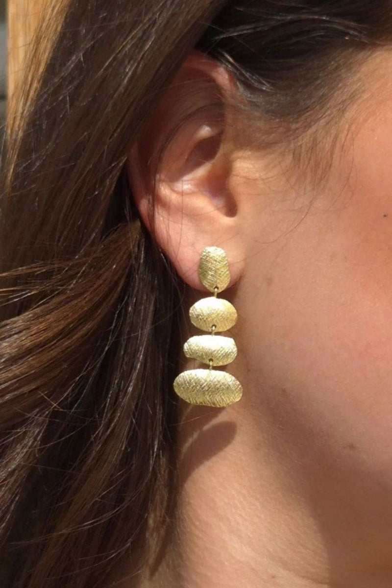 A close-up of a person's ear wearing a textured gold drop earring with three stacked gold tiered earrings