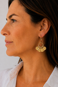 A close-up side profile of a woman wearing a gold-toned dangle earring with a circular ring and three textured coin-like discs, paired with a white collared shirt against a light neutral background.