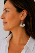 A close-up side profile of a woman wearing a dangling earring with a silver-toned ring and three clear faceted crystal drops, paired with a white collared shirt against a light neutral background.