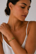 A woman wearing a delicate gold snake bracelet on her wrist, styled with a white spaghetti-strap camisole against a neutral light gray background.