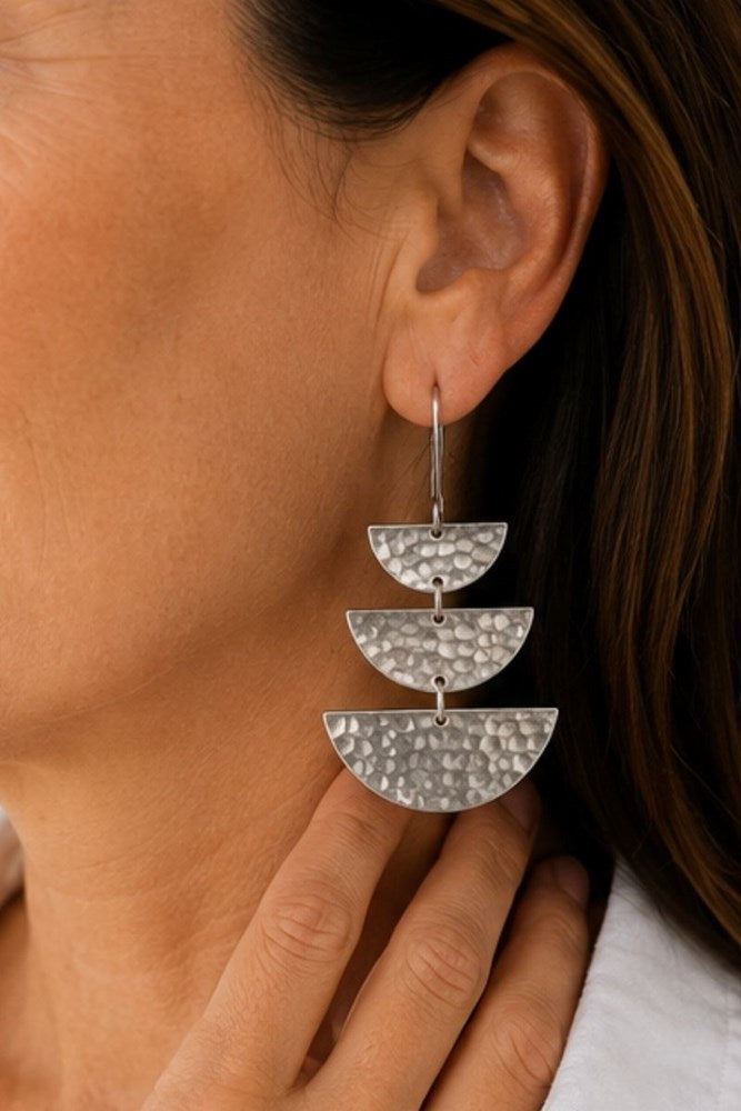 A close-up of a person wearing a silver drop earring with three textured, semi-circular tiers hanging from a single hook.