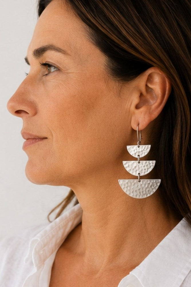 A close-up side profile of a woman wearing a pair of silver drop earrings with three stacked hammered semicircle charms.
