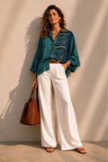 Woman wearing a blue patterned shirt and white pants, holding a brown bag against a beige wall.