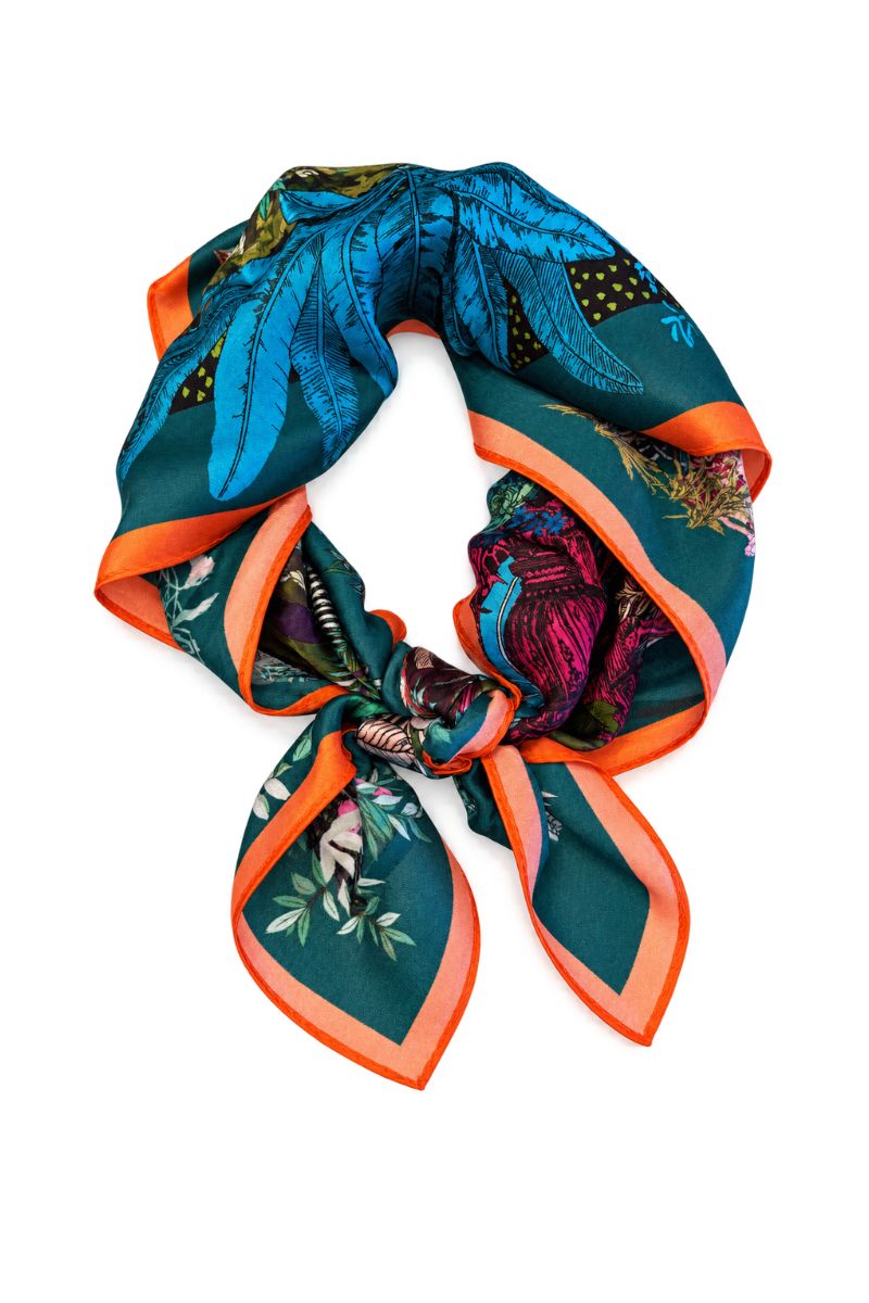A square silk scarf with a vibrant tropical print, featuring teal blue leaves, orange borders, and a dark green base, styled in a loop on a white background.