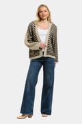 A woman modeling a beige and black patterned knit cardigan over a white top and blue jeans, standing against a light gray studio background.