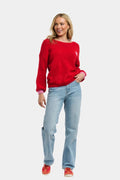 A woman modeling a red knit crewneck sweater, paired with light blue jeans and red sneakers, against a solid light gray background.