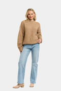 A woman modeling a light brown knit sweater and light blue jeans against a plain light gray background.