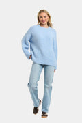 A woman modeling a light blue knit crewneck sweater and light blue jeans against a plain light gray background.