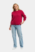 A woman modeling a bright pinkish-red knit crewneck sweater paired with light-wash blue jeans and white sneakers, standing against a light gray studio background.