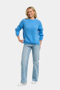 A woman modeling a bright blue crewneck knit sweater and light-wash blue jeans against a plain light gray studio background.