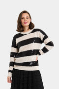 A woman wearing a black and cream striped knit sweater covered in sequins, paired with a black pleated skirt, against a light gray studio background.