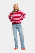 A woman modeling a pink and red horizontally striped fuzzy knit sweater, paired with light blue jeans and red sneakers, against a plain light gray background.