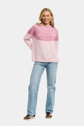 A woman modeling a chunky knit sweater with a color-blocked design featuring a textured pink yoke and a soft light pink body, paired with light blue jeans and brown sneakers, against a solid light gray background.