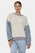 A woman modeling a color-block knit sweater with a cream ribbed body and light blue denim sleeves, paired with light-wash jeans against a plain light gray studio background.